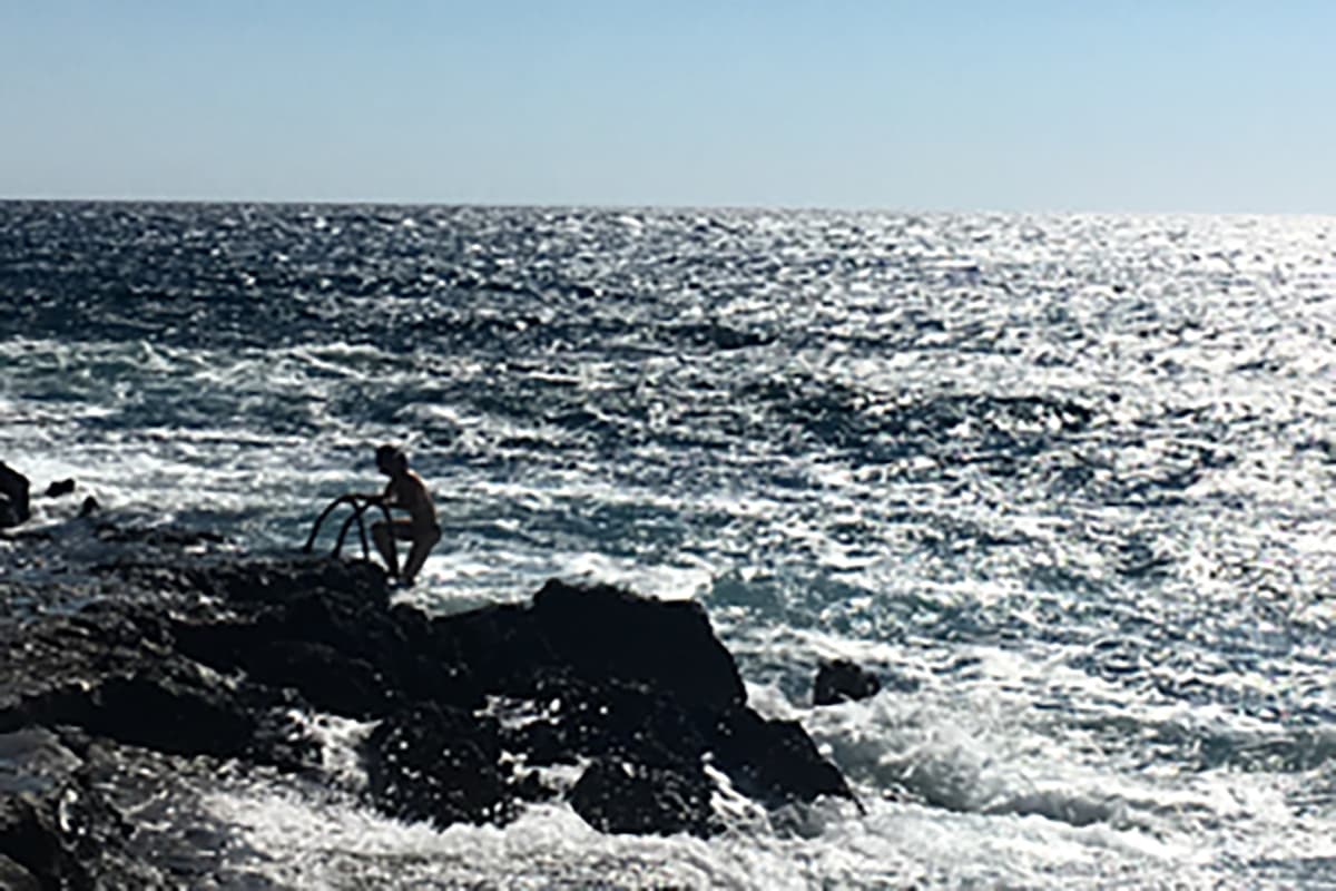 Une personne se tient sur un rivage rocheux, une échelle à la main, tandis que les vagues s'écrasent et que l'océan scintille sous un soleil éclatant.