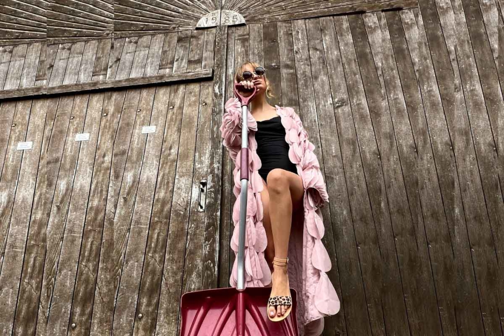 Une femme en manteau rose pose avec une pelle rose contre une porte en bois, portant des lunettes de soleil et des sandales en cuir et léopard.