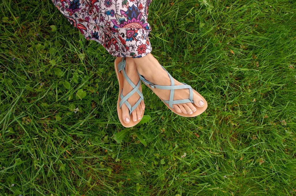 Une femme portant des sandales bleues en cuir et une robe avec des fleurs colorée est assise sur l'herbe verte.