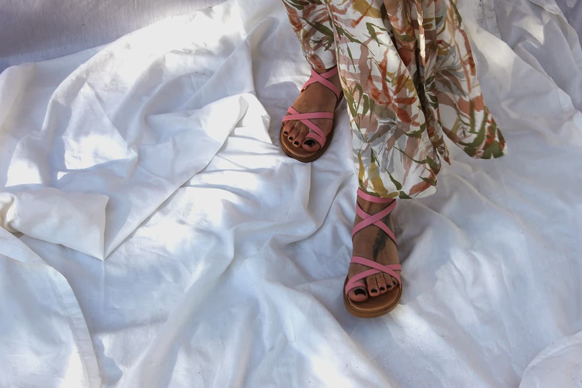 Une femme portant une robe à fleurs et des sandales à en cuir à lanières roses se tient sur un tissu blanc froissé dans une lumière douce et naturelle.