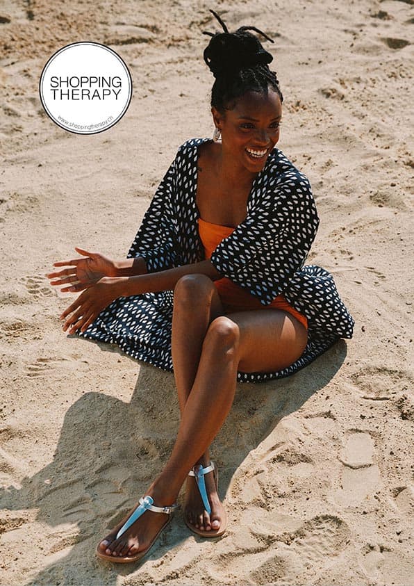 Une femme assise sur le sable, portant un des sandales argentées, un maillot de bain orange, un kimono à motifs graphiques, souriant avec ses cheveux en chignon.