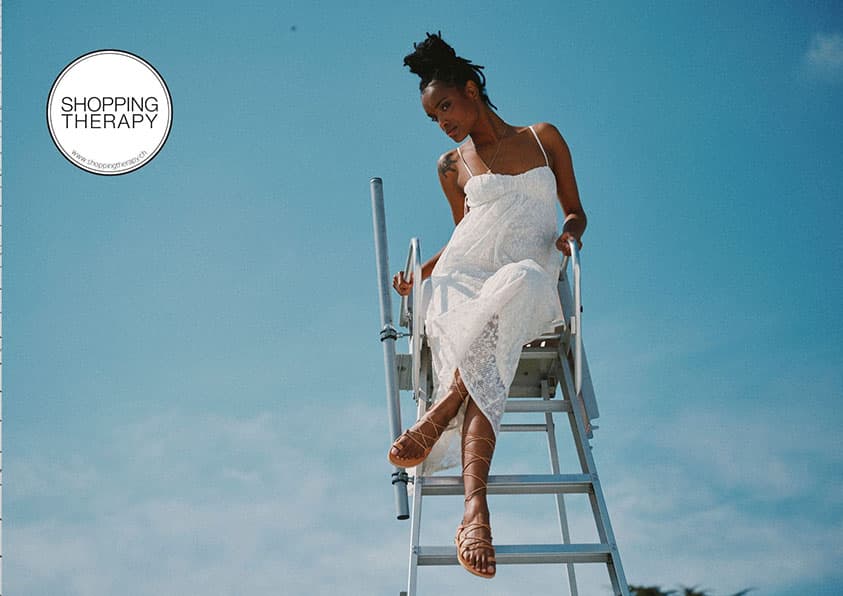 Une femme en robe blanche et sandales spartiates en cuir vachetta naturel est assise sur une chaise de maître-nageur devant un ciel bleu. Le texte « Shopping Therapy » se trouve dans le coin supérieur gauche.