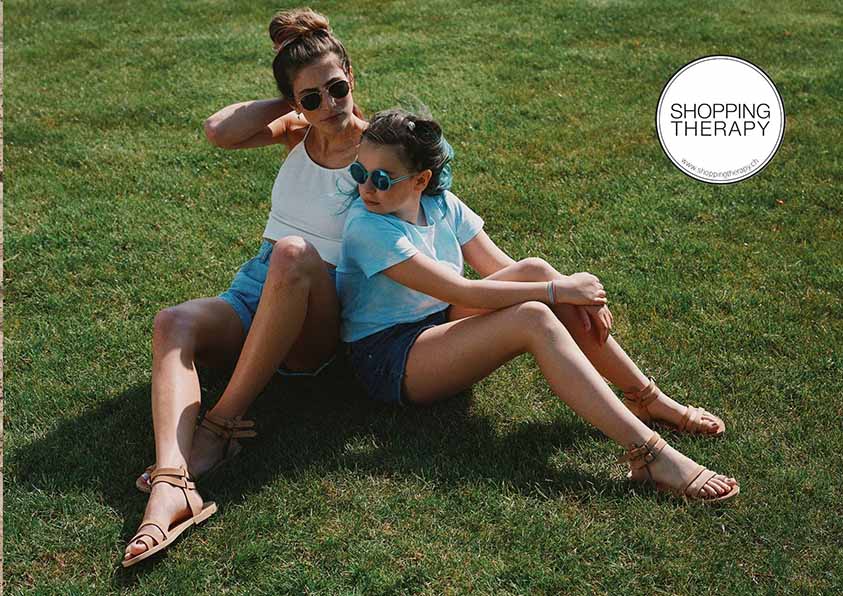 Une femme et une jeune fille sont assises sur l'herbe, vêtues dune tenue d'été décontractée des sandales spartiates en cuir et de lunettes de soleil. Le texte « Shopping Therapy » est visible dans le coin.