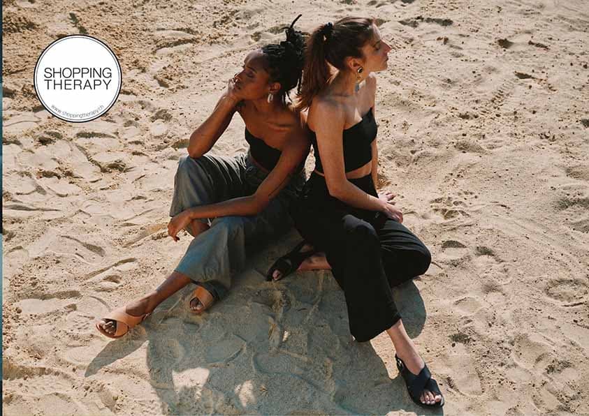 Deux femmes assises sur le sable, toutes deux vêtues de pantalons, de tops sans bretelles et des sandales croisées en cuir . Elles regardent dans des directions différentes sous le soleil. Le logo "shoppingtherapy" est en haut à gauche.