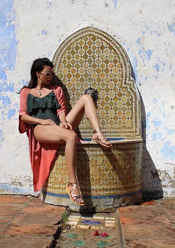 Une femme portant des lunettes de soleil, un body vert foret, un kimono orange claire et des sandales rose gold assise sur une fontaine carrelé orné contre un mur patiné blanc et bleu, avec une jambe levée, au soleil à lextérieur. Sandales Folegandros.
