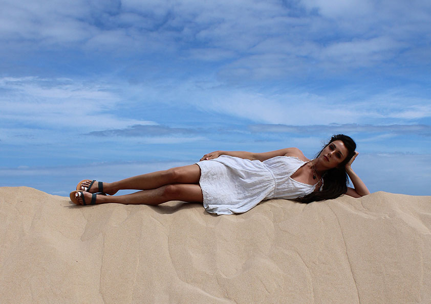 Femme en robe blanche allongée sur une dune de sable sous un ciel bleu avec des nuages, la main soutenant la tête, les jambes tendues, portant des nu-pieds en cuir gis et argenté. Sandales Epidavros.