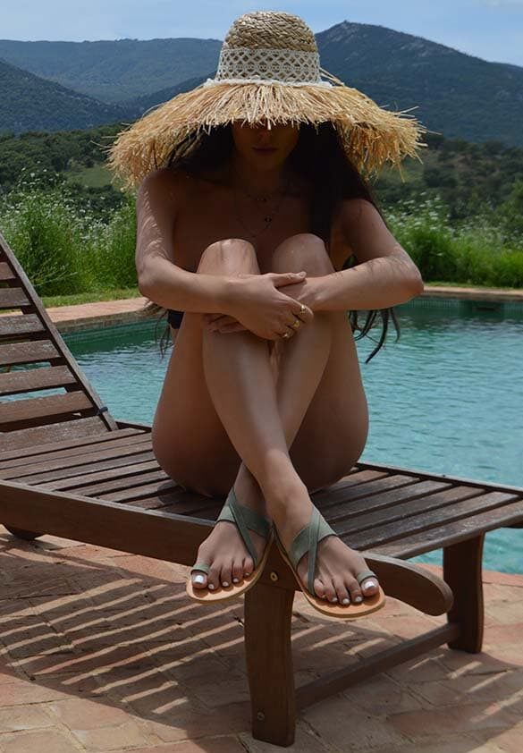 A woman sitting on a wooden deckchair by a swimming pool, wearing a large straw hat and sauge green nubuck leather sandals, with distant mountains and greenery in the background. Patmos sandals.