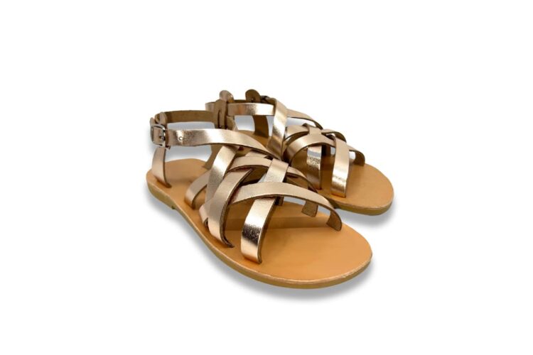 Pair of gold metallic, strappy flat sandals with adjustable ankle straps and tan soles, placed on a white background.