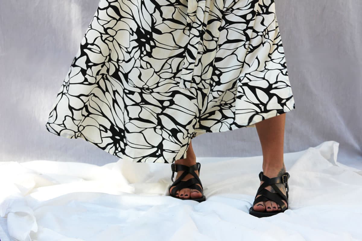 Une femme portant des sandales noires et une jupe fluide à motifs abstraits noirs et blancs se tient debout sur un drap blanc. Sandales NAXOS.