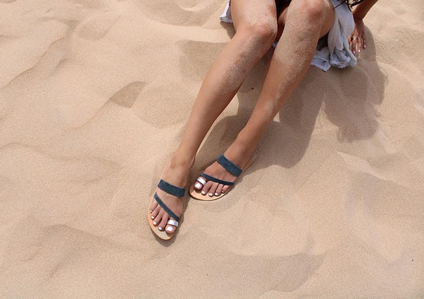 Personne assise sur une plage de sable, portant des sandales. Jambes tendues, le sable sur la peau, orteils peints en blanc. Texture de sable beige visible.